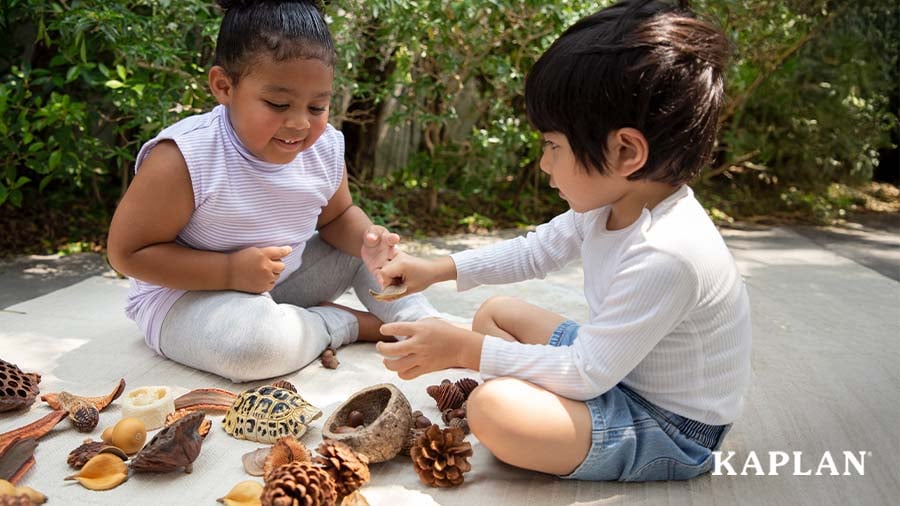 Two children play outside on a blanket with nature-inspired items such as replicas of pinecones and turtle shells