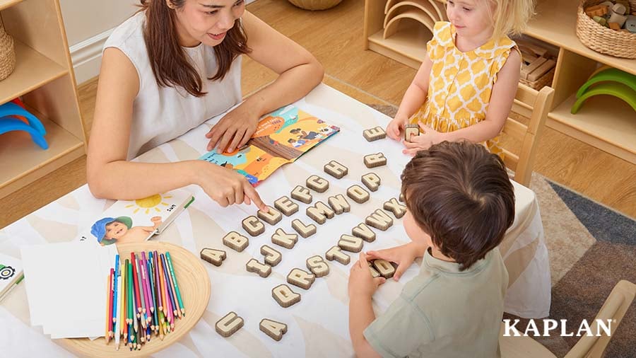 A young adult woman points out the letter "D" to two preschool-aged children as the three of them sit at a table with uppercase letters spread out on it