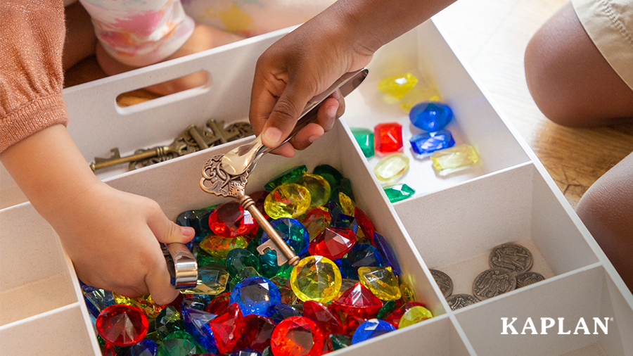 An image a a loose parts tray containing colored jewels, brass keys, faux coins, and a pair of tweezers. 