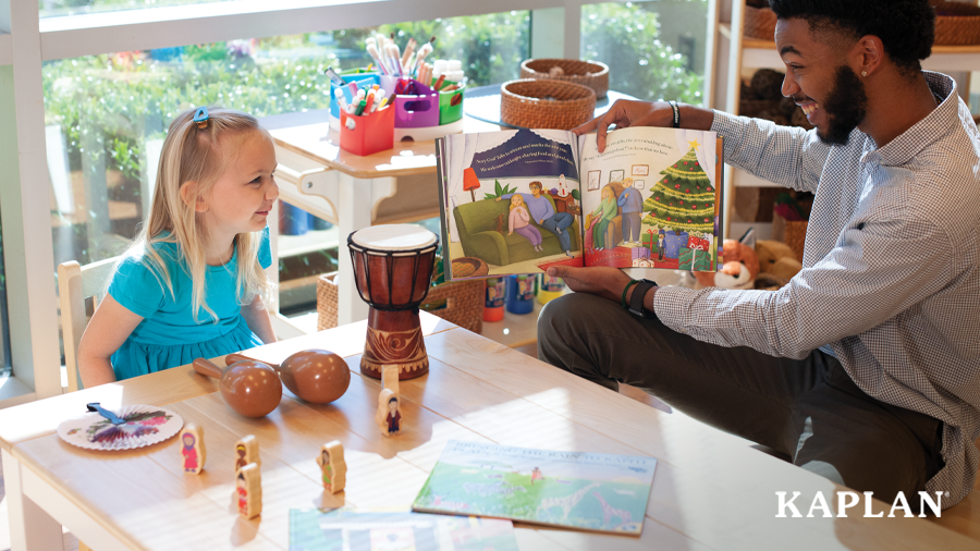 An early childhood teacher sits at a wooden chair beside a young girl in a blue dress. The teacher is holding a book open in front of the young girl. 