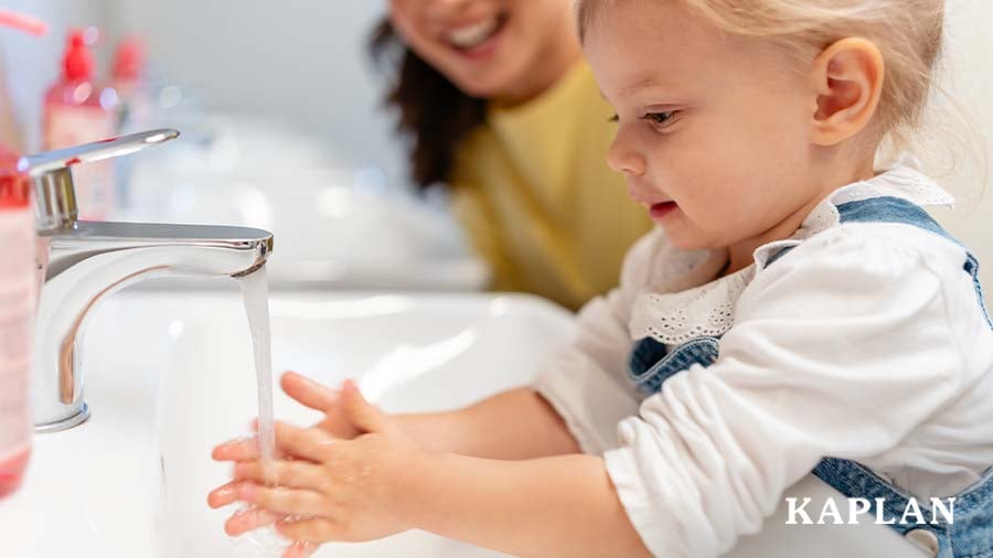 Young child washing their hands at a sink as an early educator stands close by