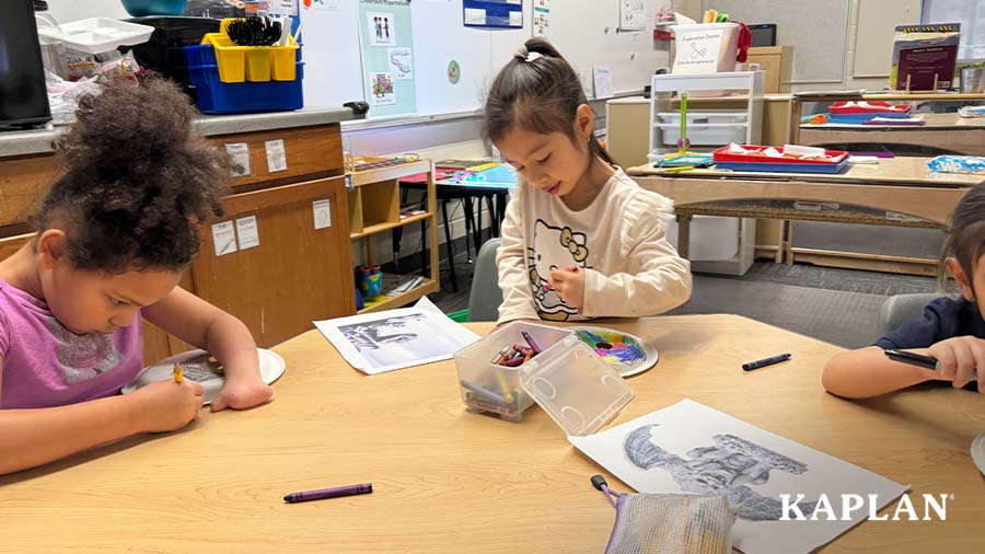 Young children stand around a wooden table, while working on an art project which requires drawing a statue face on a paper plate using crayons. 