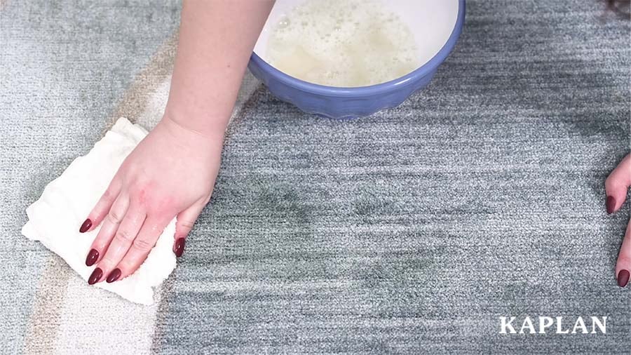 Hands holding a white cloth are blotting up a spill stain from a blue classroom rug. 