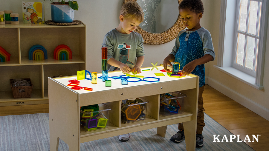 Two young boys are standing beside a light table, they are using multi colored light and color blocks to build shapes and structures on the table. 