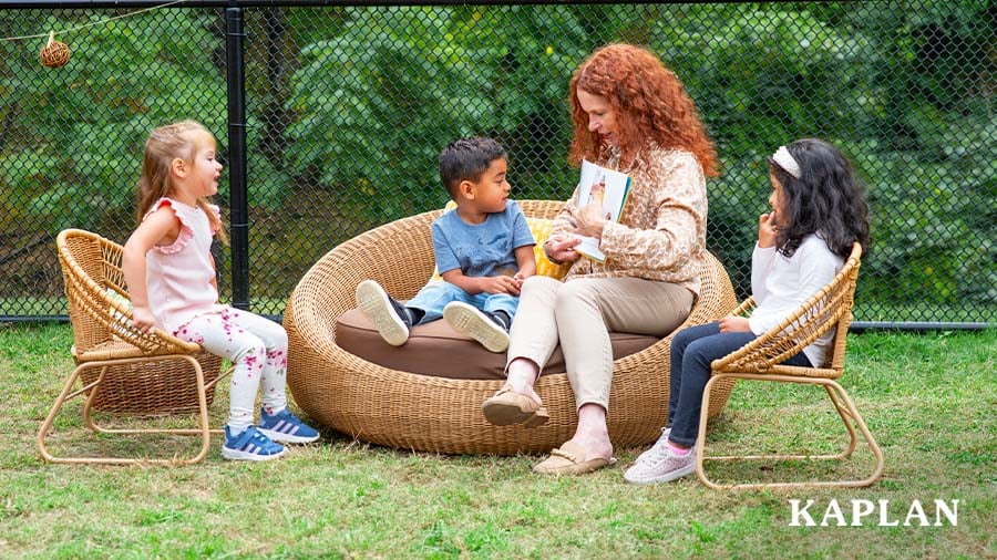Early childhood educator reads a book to three young children seated in outdoor wicker furniture