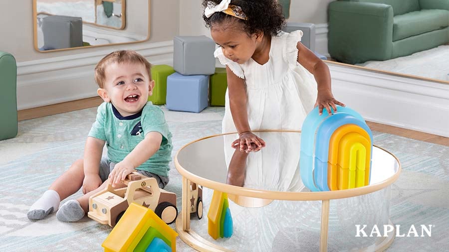 One child standing at a table while another child sits on the carpet playing with toys