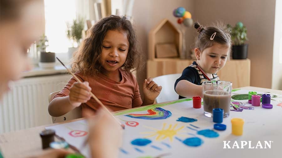 Two young children use paint to create pictures of rainbows and sunshine. 
