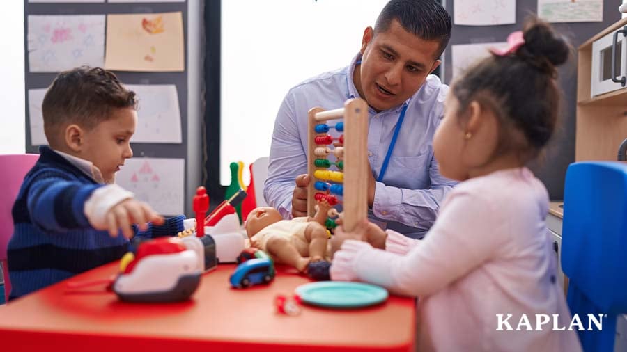 An early childhood professional sits at a table with two young children, the professional is helping one of the children move colored beads on a wooden abacus. 
