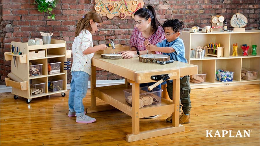 Two preschoolers in safety glasses hammer nails into wood slices at a wooden workbench while an educator guides them
