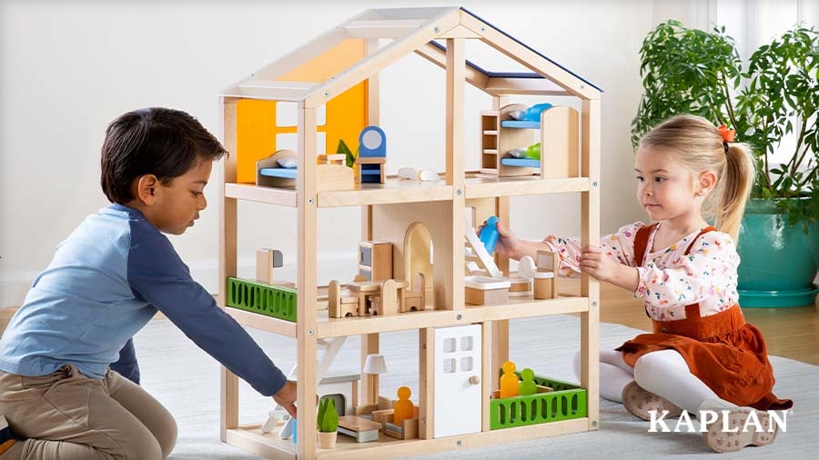 Two young children arrange tiny wooden furniture inside an open wooden dollhouse