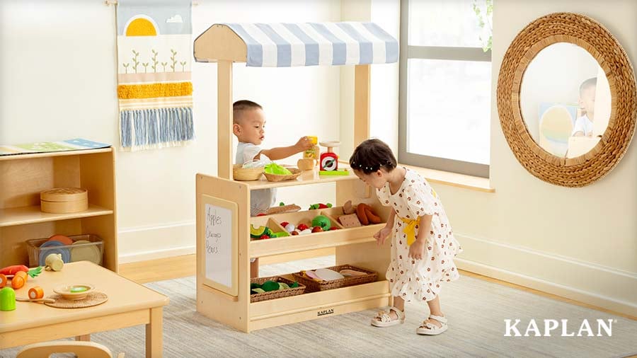 Two young children shop for pretend fruits and veggies at a wooden market stand in the dramatic play learning center