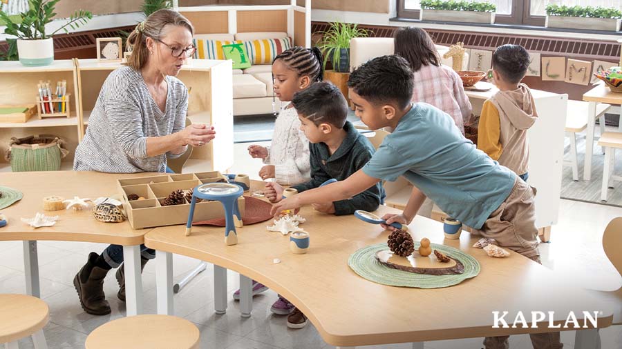 An early elementary teacher sits at a curved Sense of Place for Elementary desk, three children are also sitting at the desk. The teacher and children are looking at natural loose parts such as star fish, turtle shells, and pine cones. 