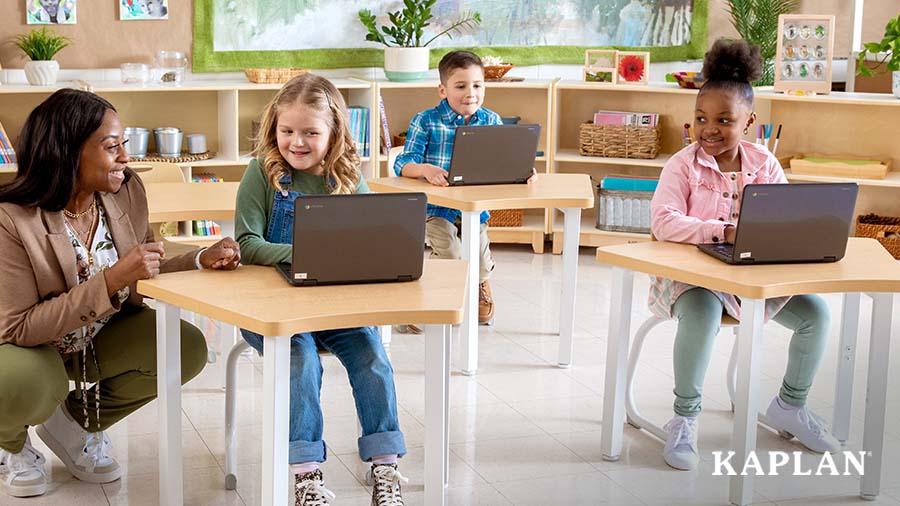 An early elementary teacher squats down beside a young girl who is sitting at a Sense of Place for Elementary desk. The girl is looking at a laptop computer which is open on the desk. 