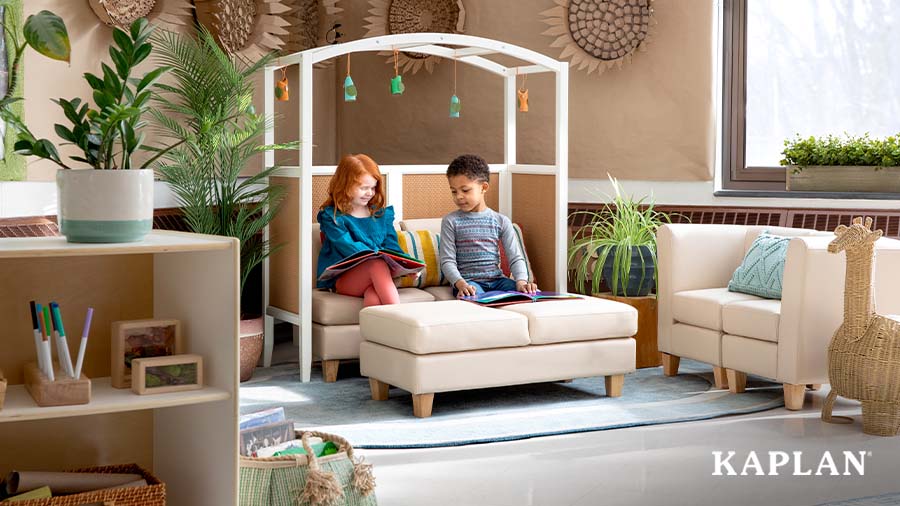 Two children are sitting on a Sense of Place for Elementary vinyl couch which is positioned underneath the Curved Arch. Both children are smiling and looking at children's books which are resting on their lap. 