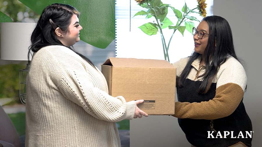 Two women are holding a brown Kaplan product box while looking at each other and smiling. 