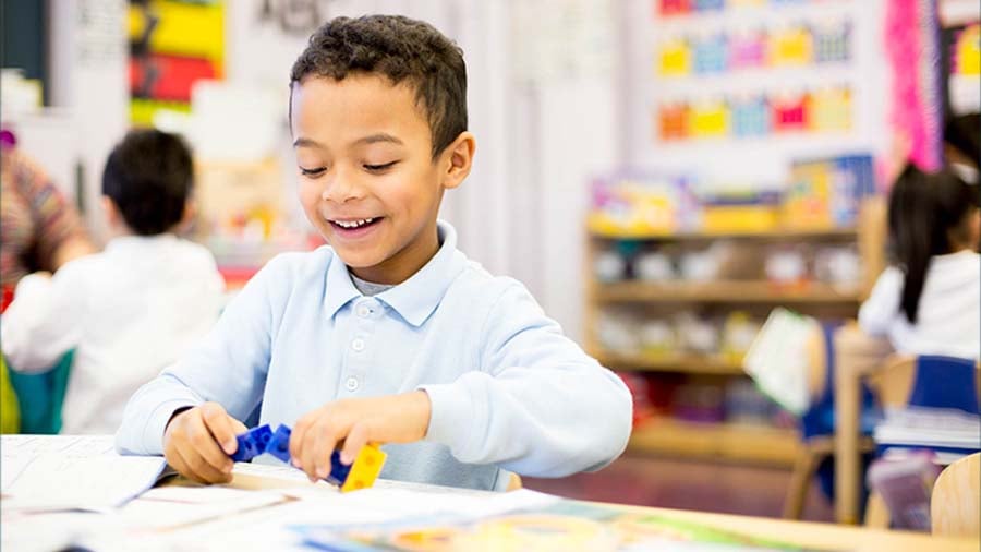 A young child sits in an early childhood classroom, the child is looking down at manipulative blocks which are in the child's hands, a smile is on their face. 