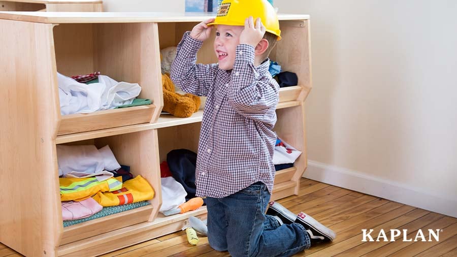 A young boy sits in a classroom dramatic play center, looking to the left of the photo, smiling while placing a construction hat on his head. 