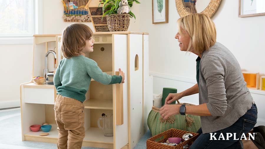 An early childhood teacher and young child smile at each other as the child opens the door on a Sense of Place for Wee Ones Sink and Refrigerator Kitchen. 