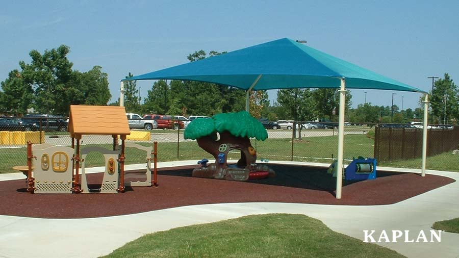 A blue sun shade covers a portion of a toddler playground area featuring a tree climbing structure and pour in place rubber mulch. 