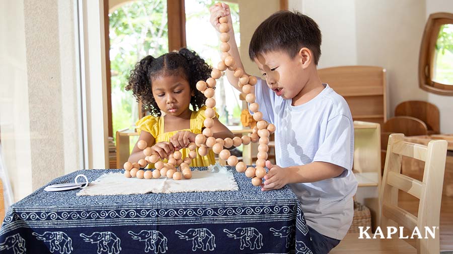 Two young children sit at a small table which is covered in a dark blue tablecloth featuring images of elephants and other shapes. The children are playing with a set of Kaplan's Natural BendiBeads.