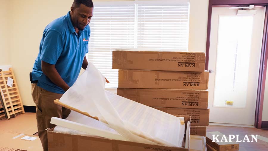 A Kaplan employee in a blue shirt stands inside an early childhood classroom, brown cardboard packaging boxes are in the room behind him. The man is opening a box and using his hands to pull a crib railing out of the box. 