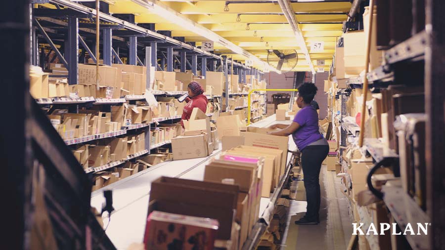Two women stand inside a packaging facility, they are looking at shelves full of brown cardboard boxes. 
