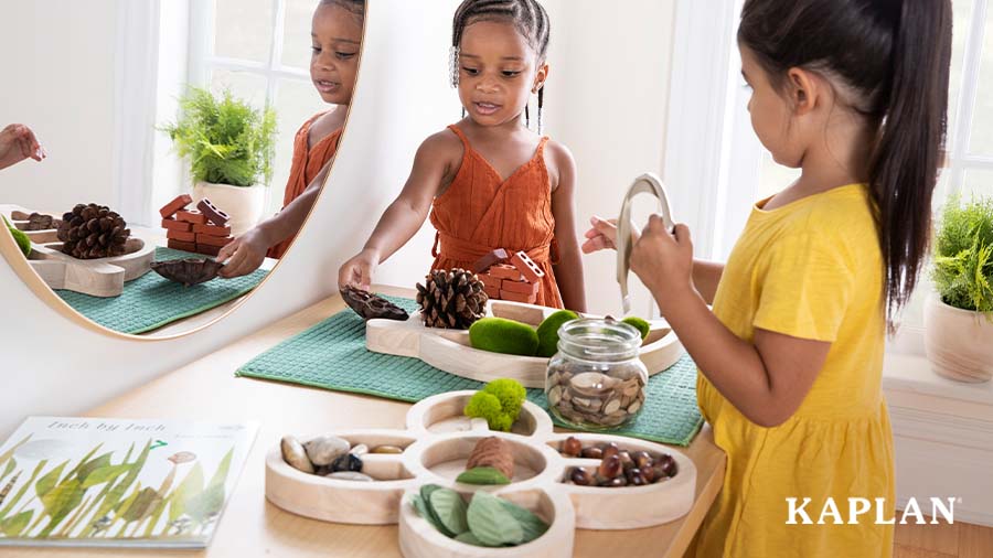 Two young children stand next to a wooden shelf with a loose parts tray and loose parts materials on top, they are both looking at the loose parts materials and reaching out as if to pick them up. 