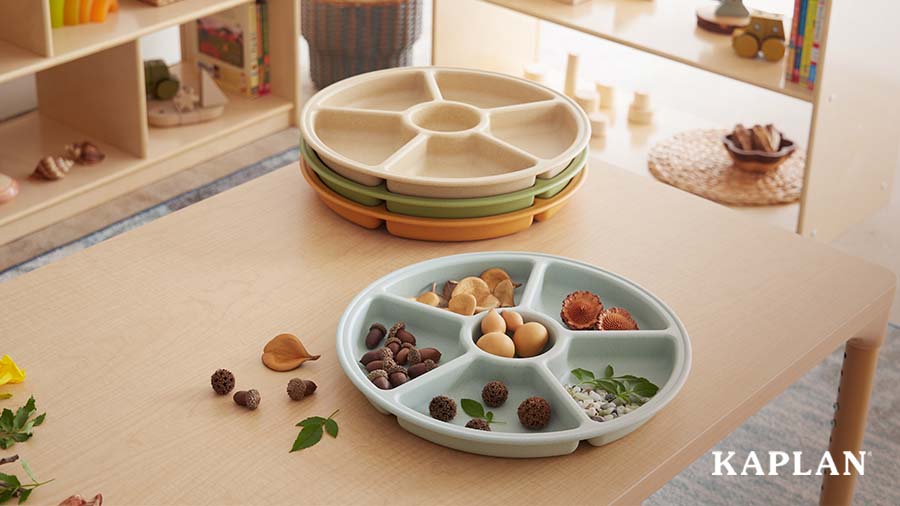 Kaplan loose parts trays sit on a wooden classroom table, there are natural materials such as acorns, leaves, and flower petals resting inside one of the loose parts trays. 