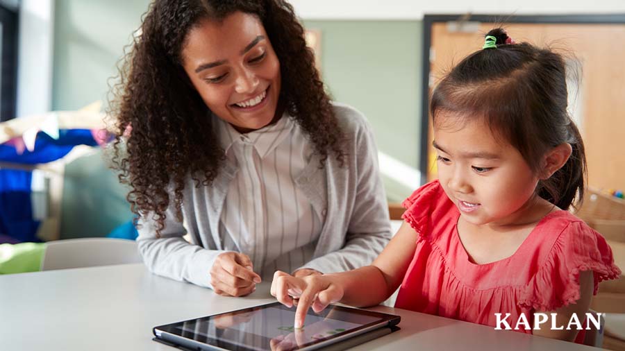 An early childhood teacher sits at a table beside a young girl in a pink shirt, the girl is looking down at a tablet on the table and is pressing her finger to the screen. 