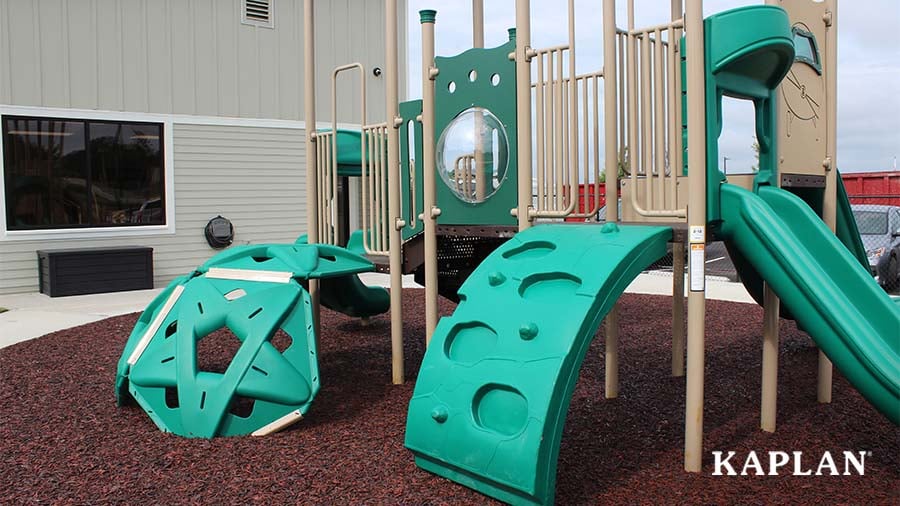 An image of a large green and tan outdoor play structure which features climbers and slides. 