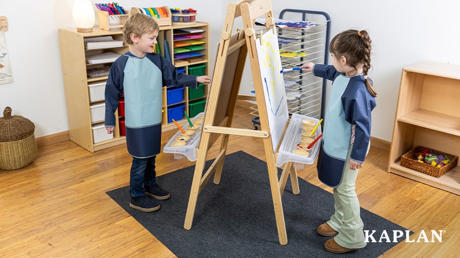 Two young children painting together at a double-sided art easel, each working on opposite sides.