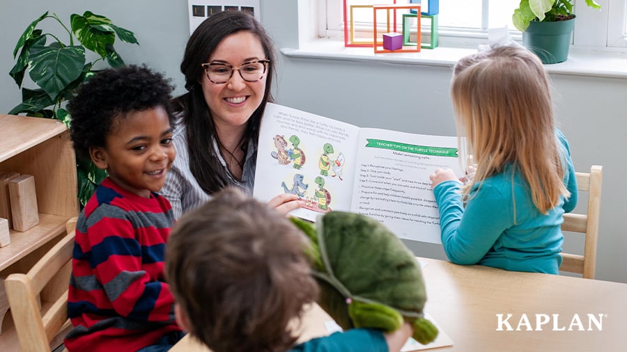 An early childhood teacher smiles while holding a book, three children sit around the teacher looking at the book and smiling. 