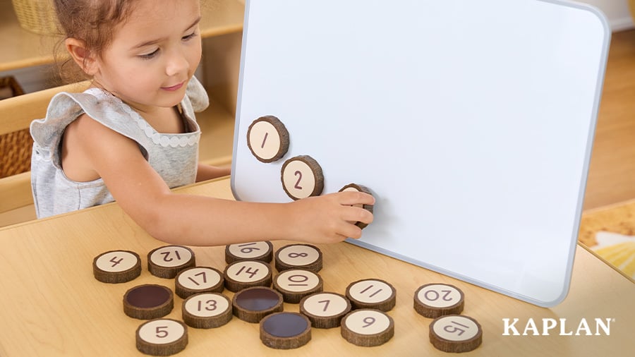 A young child sits at a classroom table, a magnetic dry erase board is on a table in front of the child. The child is placing Wooden Cookie Counters on the board in numerical order, starting with number 1. 