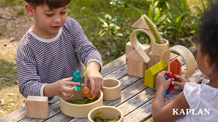 Two young children stand around a wooden table which is outdoors, the children are holding Light and Color People from Kaplan and playing with natural loose parts and wooden block shapes. 