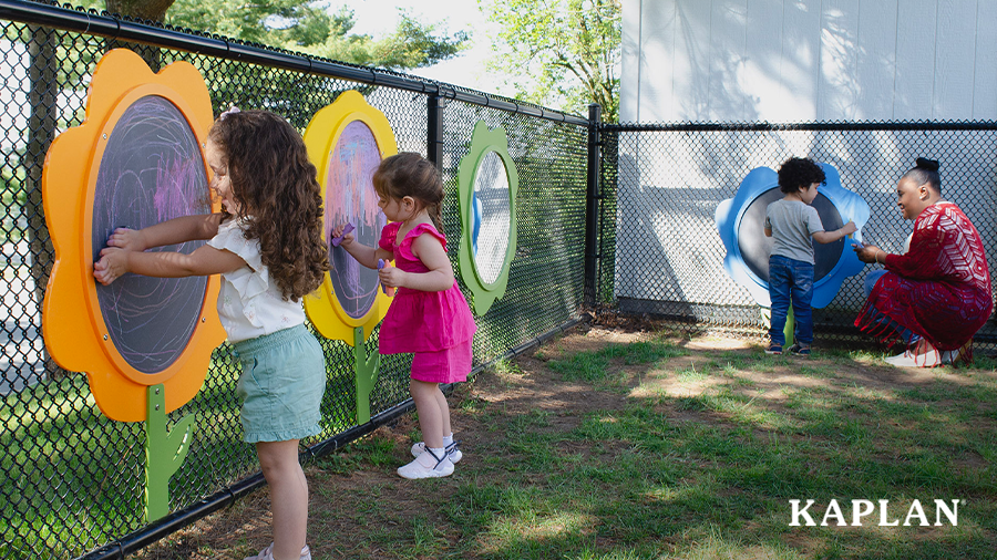 An early childhood teacher and three children are outside on the playground, the children are drawing on Kaplan's Wooden Fence Easels which are attached to a chain-link fence. 