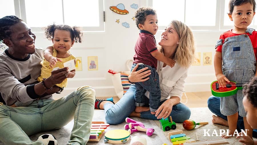 Two early childhood educators sit on the floor as they interact and play with a group of toddlers. 