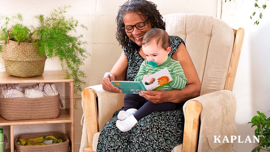 An early childhood teacher sits in a classroom glider, smiling as she reads a book to a young child seated on her lap. 