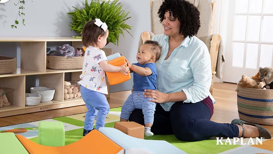 An early childhood educator sits on the floor with two infants, one on her lap, the other standing in front of them holding a soft foam block. 