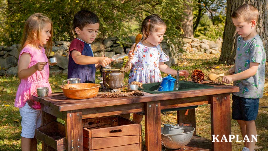 A group of four young children stand at an outdoor mud kitchen, they each have a kitchen utensil in their hands and are pouring pinecones and flowers into a large clay pot. 