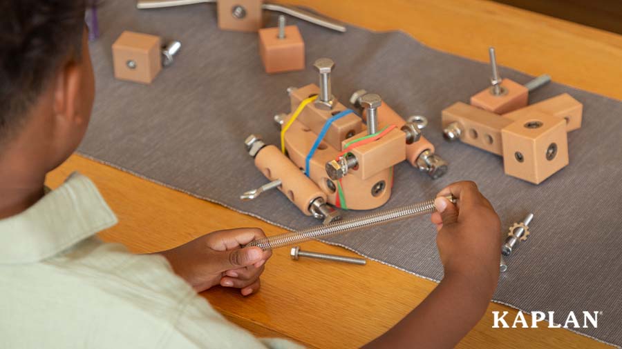 A young child holds a metal spring in their hand while looking down at a pile of construction loose parts. 