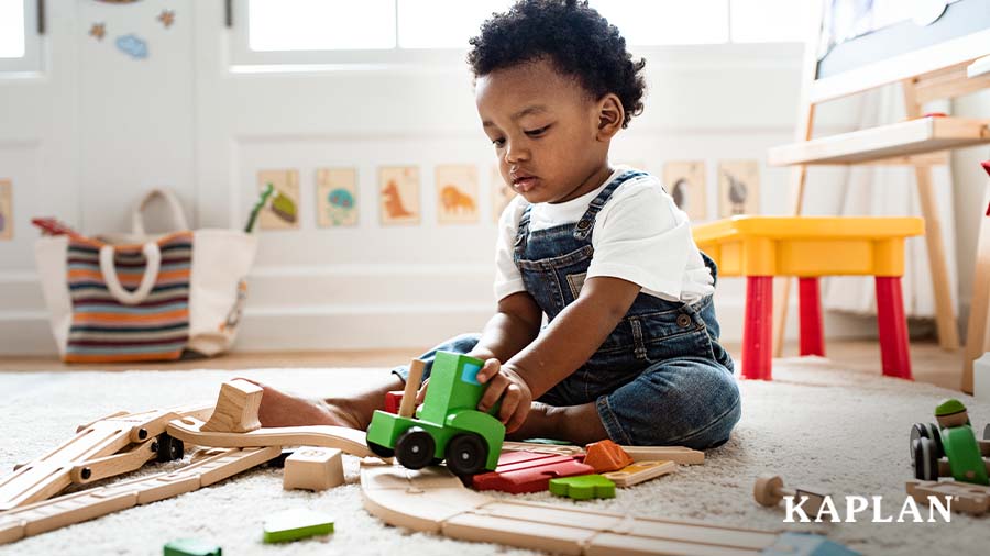 An infant sits on a carpeted play area in a home-based child care facility, he is looking down at a wooden train in his left hand. 