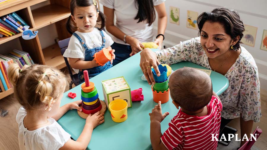 An early childhood teacher sits at a blue table, there are three young children at the table. The teacher and children are stacking cups and wooden blocks. 