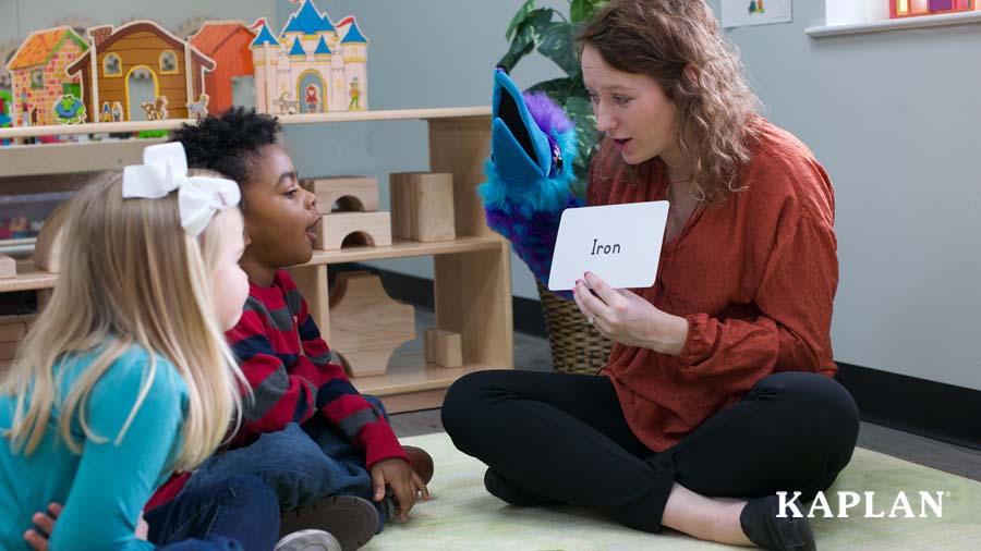 An early childhood teacher sits on a classroom carpet, holding a word card up that shows the word "Iron." Two children sit on the carpet in front of the teacher, looking at the word card. 