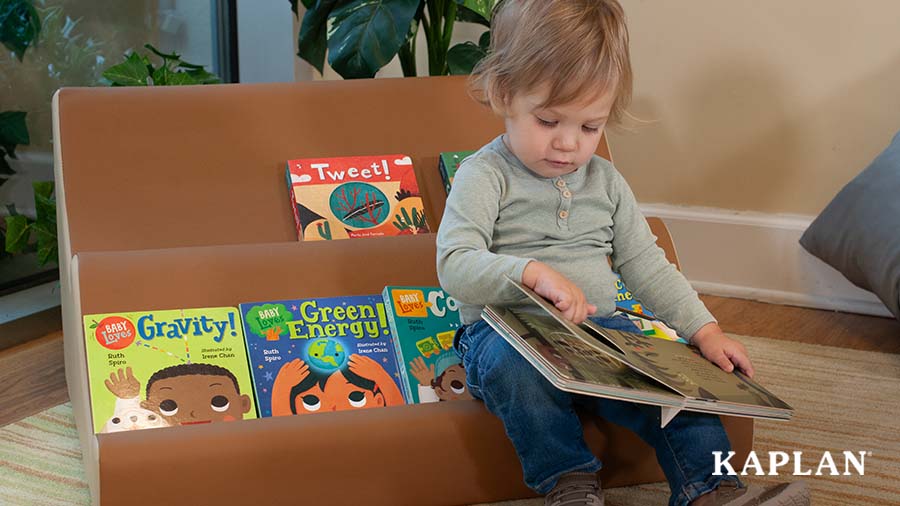 A young child sits in front of a wooden bookshelf, an open book across its lap while the child looks down at the open pages. 