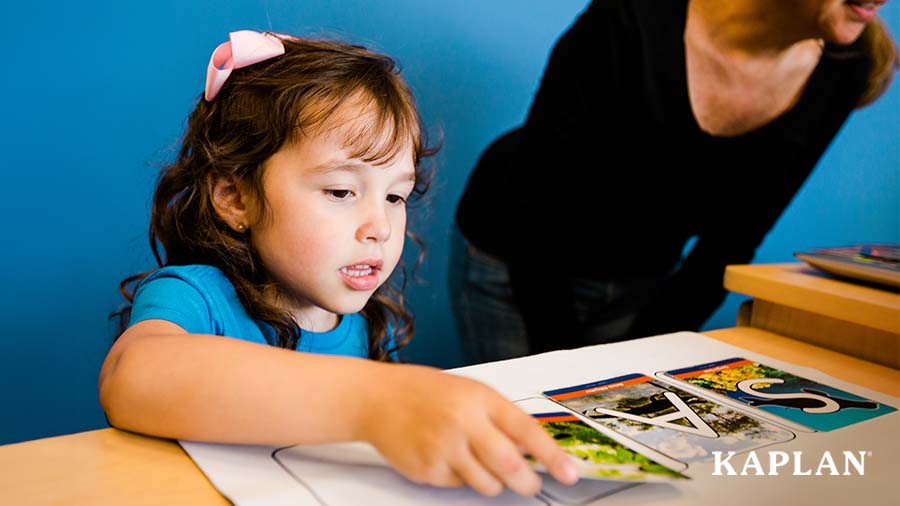 A young girl in a blue shirt sits at a wooden table, placing a letter card on a piece of white paper. 