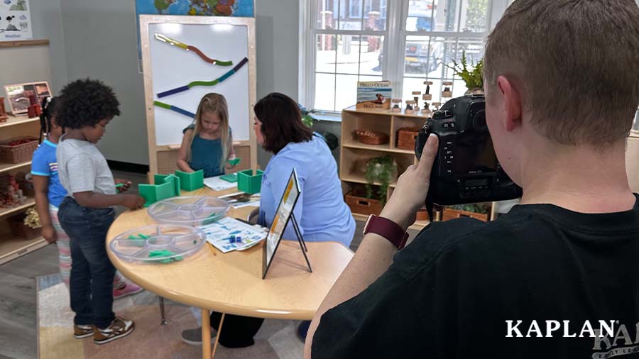 An image showing behind the scenes of the Kaplan Model Call. The Kaplan photographer is taking a picture of young children and an early childhood teacher working on an art activity at a wooden classroom table. 