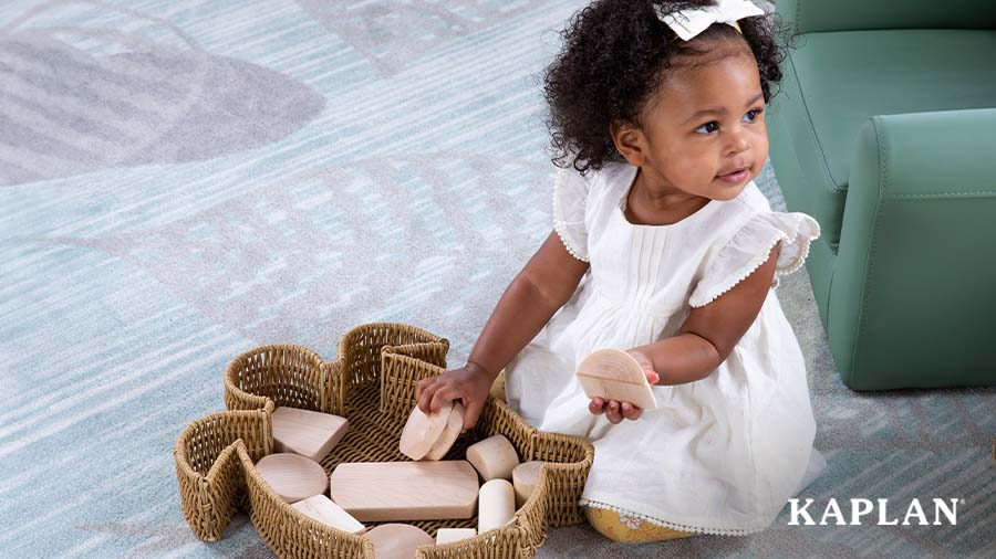 A young toddler sits on a Sense of Place carpet, in front of her is a Sense of Place basket with wooden blocks. She is holding the blocks in her hands while looking over her left shoulder. 