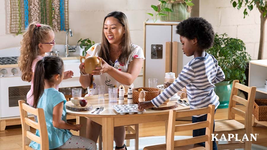 A young adult female has pretend-play mealtime with three preschool-aged children at a wooden oval table