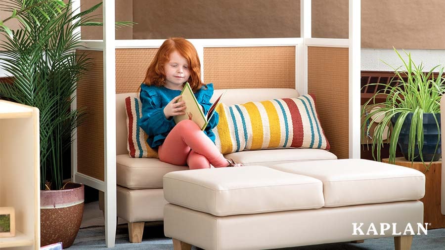 A young girl of early-elementary age looks at a picture book on a soft chair underneath a classroom arch