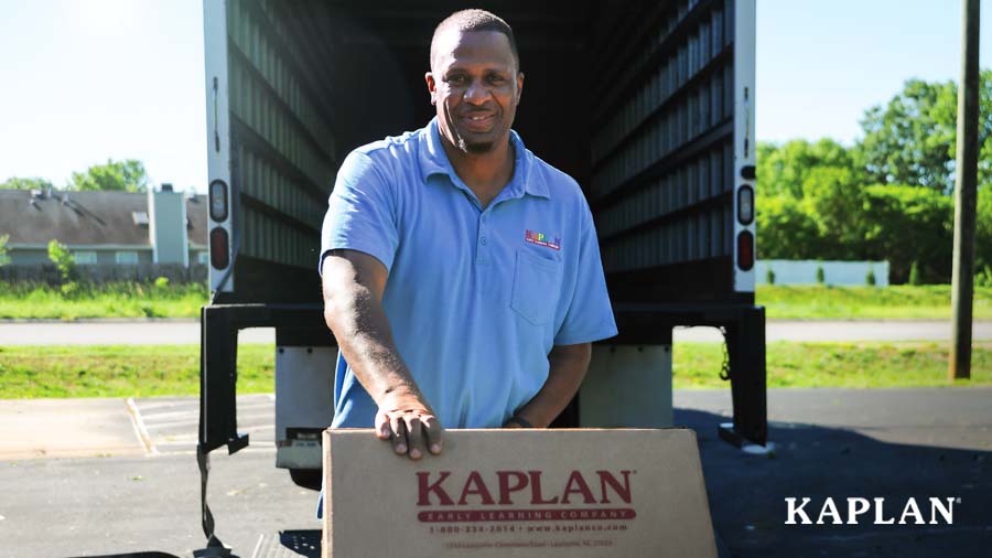 A Kaplan employee in a blue shirt stands behind an open delivery truck, he has a brown package in his hands that says "Kaplan Early Learning Company."